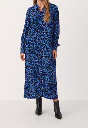 Woman wearing a long blue and black leopard print button-up dress with long sleeves and black ankle boots, standing on white floor.