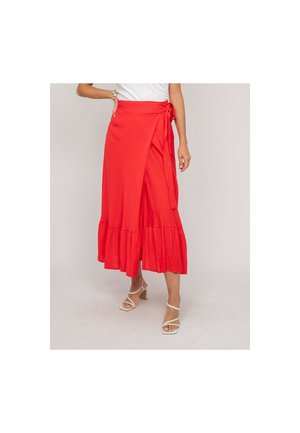 Femme portant une jupe portefeuille rouge à ourlet volanté et des sandales blanches à talons avec lanières, debout devant un fond clair uni.