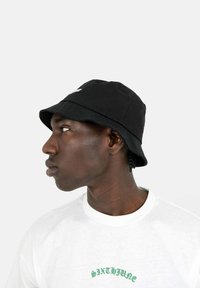 Cappello a secchiello nero in tessuto di cotone con un ampio bordo flessibile. Presenta ricami bianchi sul davanti. Indossato con una maglietta bianca.