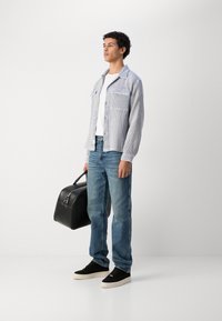 Blå og hvit stripete skjorte med krage, hvit t-skjorte, lyseblå jeans, svarte joggesko, holder en svart lær treningsbag.