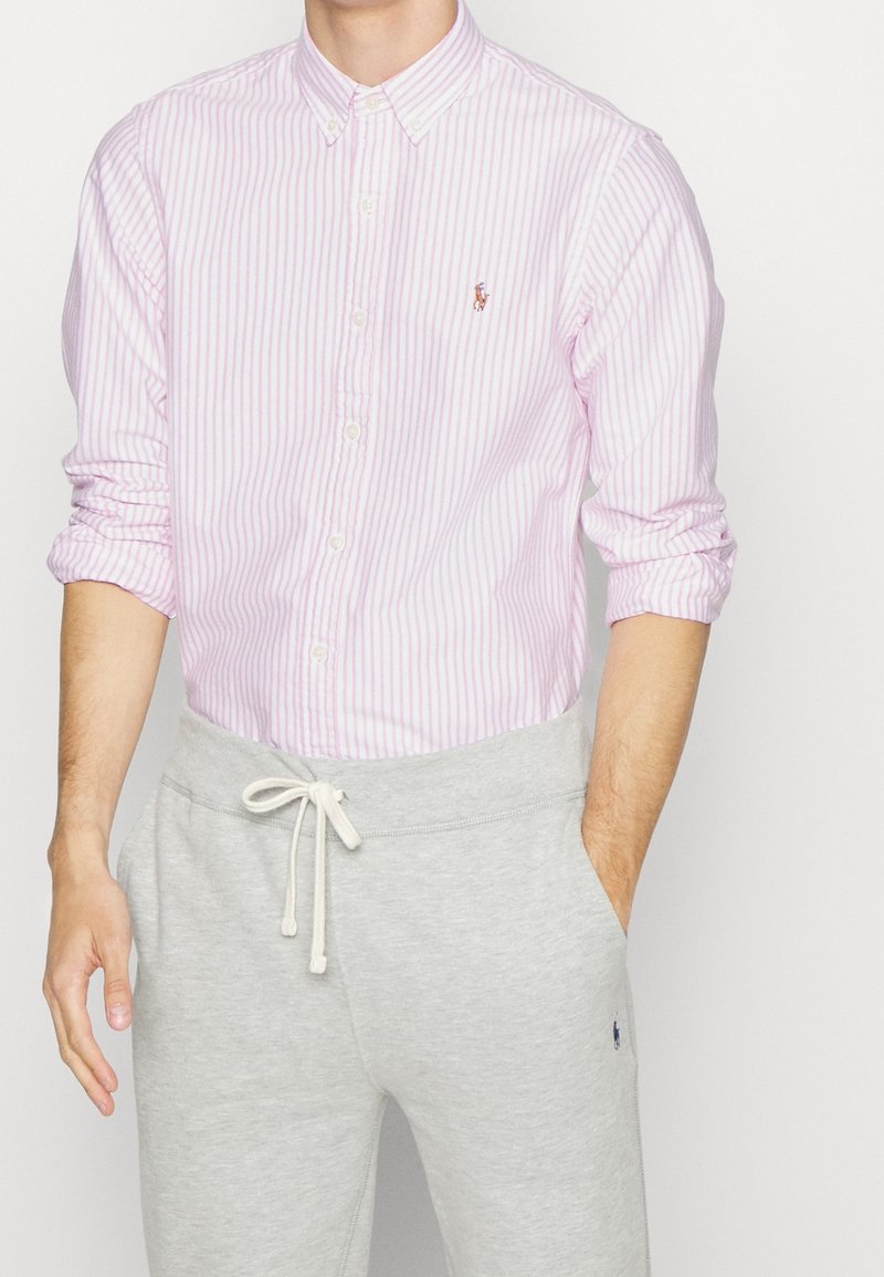 Světle růžová a bílá pruhovaná košile na knoflíky s límečkem, v kombinaci se šedými tepláky s šňůrkou a bočními kapsami.