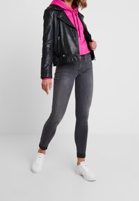 Chaqueta de motociclista de cuero negro sobre una sudadera rosa brillante, combinada con jeans ajustados grises y zapatillas de plataforma blancas. Texturas suaves y diseño ajustado.