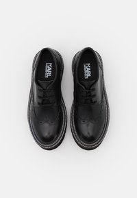 Chaussures en cuir noir avec des détails en brogue, design à lacets, surpiqûres blanches contrastantes et bout arrondi. Logo de la marque visible à l'intérieur.