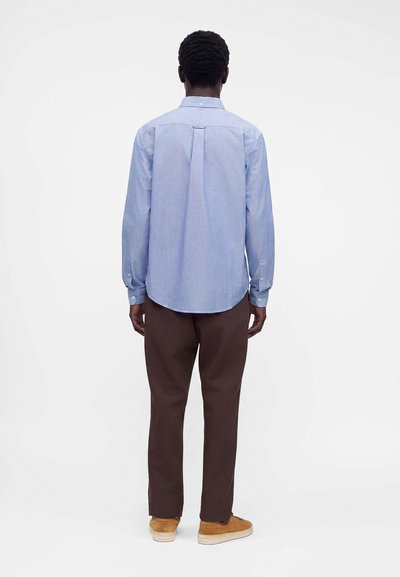 Homme debout, dos tourné, portant une chemise bleu clair à manches longues, un pantalon marron foncé et des baskets beige sur un fond blanc uni.