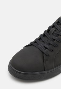 Svart syntetisk sneakers med en slät matt yta, platta snören, rund tå och en strukturerad gummisula med ett subtilt mönster.