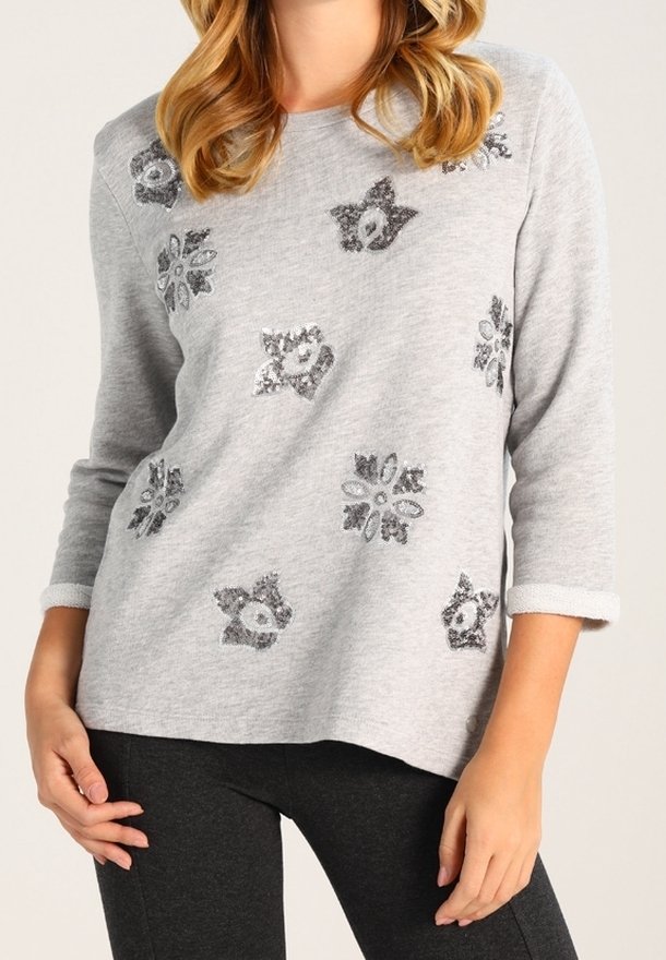 Pull gris avec manches trois-quarts, orné de fleurs en sequins argentés. Texture douce et coupe décontractée. Porté avec des leggings noirs.