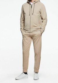 Sudadera beige con cremallera y cordones, joggers ajustados a juego y zapatillas blancas. Tejido suave con una textura lisa y sin patrones.