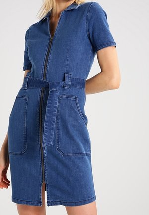 Une femme porte une robe en jean bleu à manches courtes avec une fermeture éclair à l'avant, une taille ceinturée et de grandes poches devant, debout devant un fond uni.
