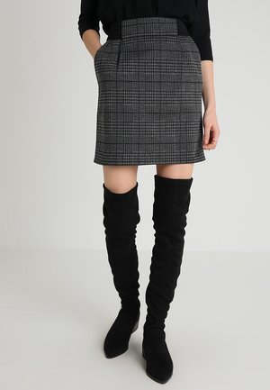 Femme portant une jupe grise à carreaux avec poches et des bottes noires par-dessus le genou, debout devant un fond clair uni.