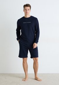 Mies seisoo paljain jaloin, päällä tummansininen Tommy Hilfiger -neule ja samansävyiset shortsit, taustalla yksinkertainen valkoinen seinä.
