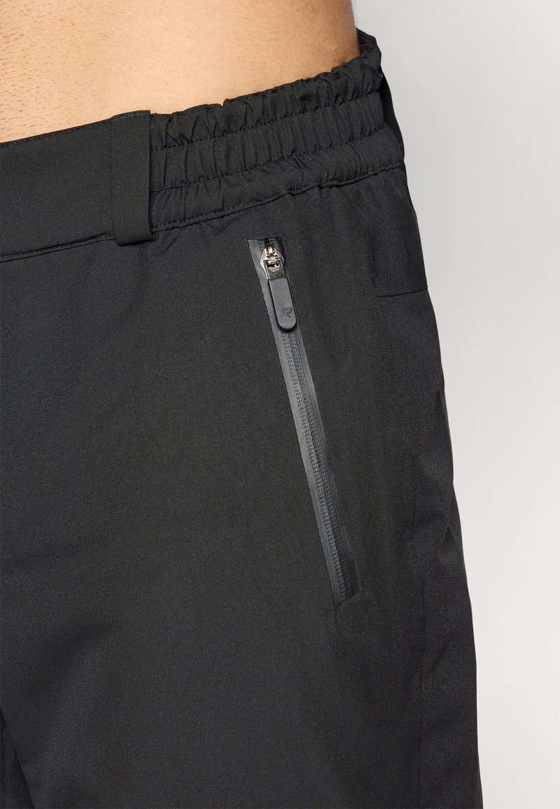 Close-up van een persoon die zwarte broek met een elastische tailleband en een verticale ritszak aan de rechterzijde draagt.