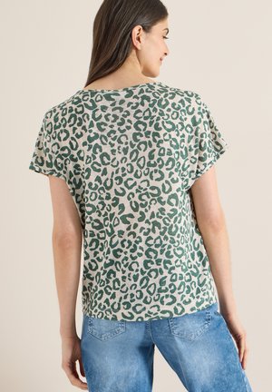 Femme aux longs cheveux bruns portant un haut beige à manches courtes avec un imprimé léopard vert et un jean bleu clair, vue de dos sur un fond uni.