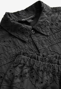 Sort bluse lavet af tekstureret stof med blomsterbroderi og krave. Inkluderer matchende elastisk taljebånd.