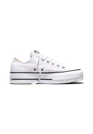 Zapatilla baja de lona blanca con cordones blancos, franja negra en la suela y puntera de goma blanca texturizada.