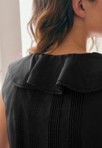 Schwarze Bluse mit gerafftem Kragen und zarter Spitzenbordüre, mit vertikalen Streifen und glatter Textur.