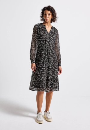 Woman wearing a black knee-length dress with white abstract patterns, long sheer sleeves, and white sneakers, standing on a white background.