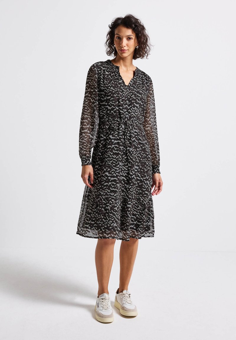 Femme portant une robe noire midi avec des motifs abstraits blancs, des manches longues en voile et des sneakers blanches, se tenant sur fond blanc.