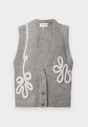 Personne noire portant un gilet en tricot gris sans manches avec des motifs blancs, une jupe noire taille haute avec une fente à l'avant, et des bottines marron.