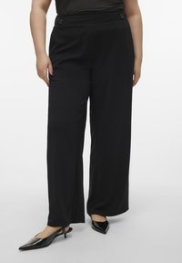 Pantalons noirs à largeur large en tissu lisse avec une taille haute, ornés de deux accents de boutons. Associés à des escarpins noirs à bout pointu.