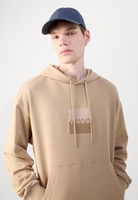 Felpa beige con tasca frontale e logo HUGO in un design quadrato; presenta lacci e una texture leggera. Cappellino da baseball blu navy con motivo rigato.
