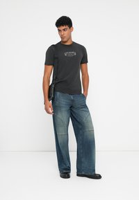 Camiseta de algodón negra con logo impreso, combinada con jeans de pierna ancha azules con detalles de parches y una textura suave. Bolso negro de hombro.