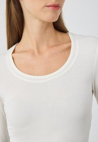 Camicia bianca a maniche lunghe con scollatura rotonda, realizzata in un tessuto leggero e fluido, dal design aderente. Dettagli di cucitura discreti sul collo.