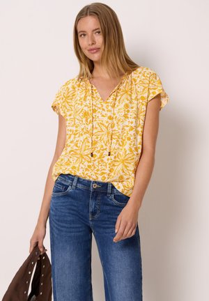 Jeune femme portant un chemisier jaune à fleurs à manches courtes et un jean bleu, tenant une veste marron dans sa main gauche.