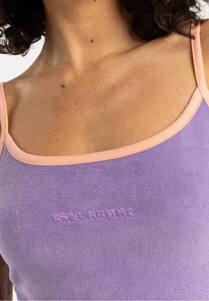 Frau, die ein lilafarbenes Tanktop mit pfirsichfarbenem Besatz und erhabenem Text in der Mitte der Brust trägt, wobei der Hals- und obere Brustbereich sichtbar ist.