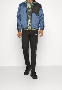 Blauer und schwarzer Windbreaker mit elastischen Bündchen, gerafftem Saum und gelben Akzenten, kombiniert mit schwarzen, enganliegenden Hosen und schwarzen Sportschuhen.