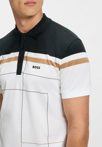 Vit polo-shirt med svarta och guld horisontella ränder, svart krage, tre-knapps knappslå och varumärkeslogotyp på bröstet.