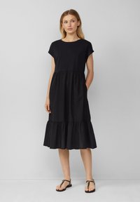 Schwarzes Baumwollkleid mit kurzen Ärmeln, Rundhalsausschnitt und fransigem Saum. Verfügt über einen gestuften Rock und seitliche Taschen. Trägt man mit flachen Sandalen.