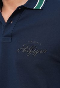 Navy polo shirt made of textured fabric, featuring a contrasting green and white striped collar and an embroidered black logo.