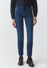 Tumedat sinised teksapüksid slim-fit kujunduses, viie tasku ja peente õmbluse detailidega. Omab klassikalist kõrget taljet ja kitsenevaid jalgu.