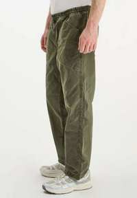 Man in olijfgroene ontspannen broek met elastische tailleband en witte sportschoenen, staand tegen een effen achtergrond.