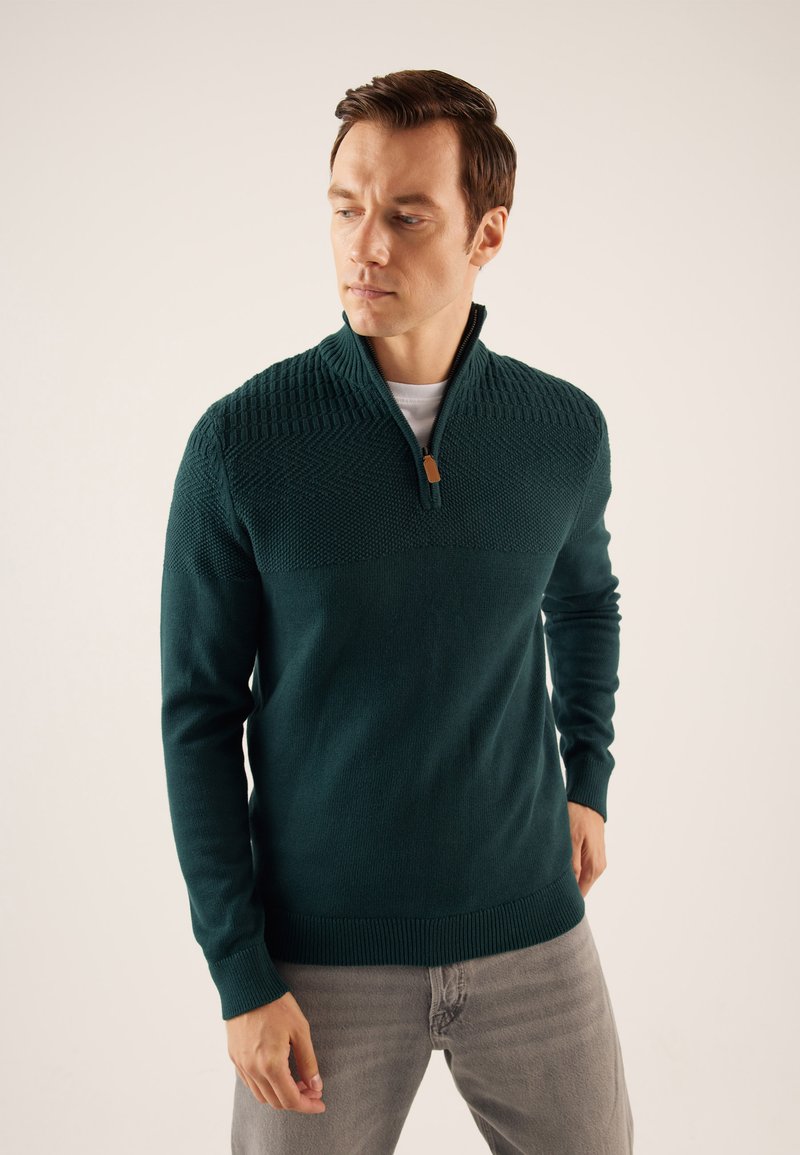 Dunkeltürkiser Pullover mit hohem Kragen und halbem Reißverschluss. Strukturiertes Strickdesign über der Brust, gerippte Bündchen und Saum. Über einem weißen Hemd getragen.