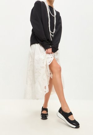 Schwarzes Sweatshirt mit langen Ärmeln, ein weißer Spitzenrock mit gerafftem Saum und schwarze Schuhe mit einer weißen Sohle und Riemen-Detail. Perlenkette als Akzent.