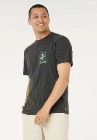 Maglietta nera in cotone con maniche corte, scollo tondo e una grafica turchese con palme sul lato sinistro del petto. Sfondo bianco.