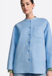 Jatak iz svetlo modrega volnenega mešanice z okroglim ovratnikom, zapenjanjem na gumbe spredaj, dvema velikima žepoma in dolgimi rokavi. Detajl teksturirane površine.