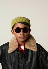 Veste en cuir noir avec un col en fausse fourrure beige, chemise rouge en dessous, lunettes de soleil noires rondes et un bonnet en laine vert.