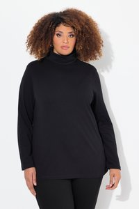 Donna con capelli ricci che indossa un top nero a collo alto e maniche lunghe e pantaloni neri, in piedi su uno sfondo bianco.