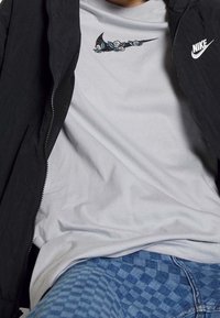Grå Nike-t-shirt med blommönster ovanför loggan, ihopparad med en svart zip-up-jacka och blå rutiga jeans.