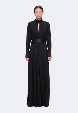 Femme debout portant une longue robe noire avec des manches longues transparentes, un col montant, une fente à l'avant et une large ceinture.
