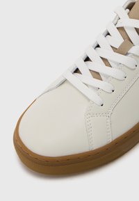 Nærbilde av hvite lær sneakers med hvite snøringer og brun gummisåle, som viser detaljert søm og skoens tunge.