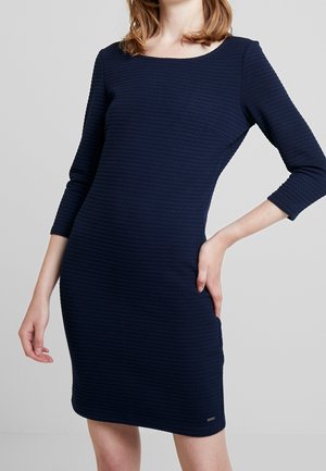 Frau trägt ein eng anliegendes, strukturiertes marineblaues Kleid mit Dreiviertelärmeln vor einem schlichten hellgrauen Hintergrund.
