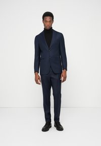 Marineblauer Anzug mit tailliertem Blazer, Ein-Knopf-Verschluss und passenden Hosen. Kombiniert mit einem schwarzen Rollkragenpullover und schwarzen Schuhen.