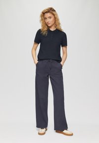 Marineblauwe polo met knopen en korte mouwen, gecombineerd met een brede, donkere broek. Model draagt witte en lichtbruine sneakers.