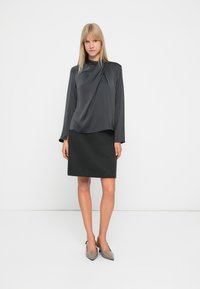 Blusa grigia scura a maniche lunghe con design drappeggiato abbinata a una gonna grigia scura aderente. La modella indossa tacchi a punta grigi chiari.