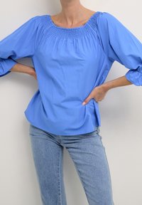 Blouse bleue avec un décolleté carré, des détails de smocks froncés et des manches bouffantes trois-quarts ; associée à un jean en denim bleu clair.