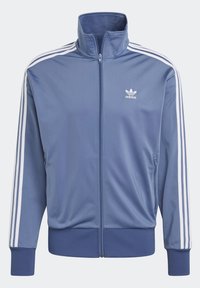 Sininen urheilutakki, jossa on korkea kaulus, täyspitkä vetoketju edessä, valkoiset kolmen raidat -koristelut hihoissa sekä resorit hihansuissa ja helmassa.