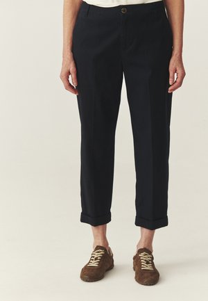 Osoba ubrana w czarne, krótkie spodnie z podwiniętymi nogawkami i brązowe zamszowe trampki z beżowymi sznurowadłami, stojąca na jednolitym tle.